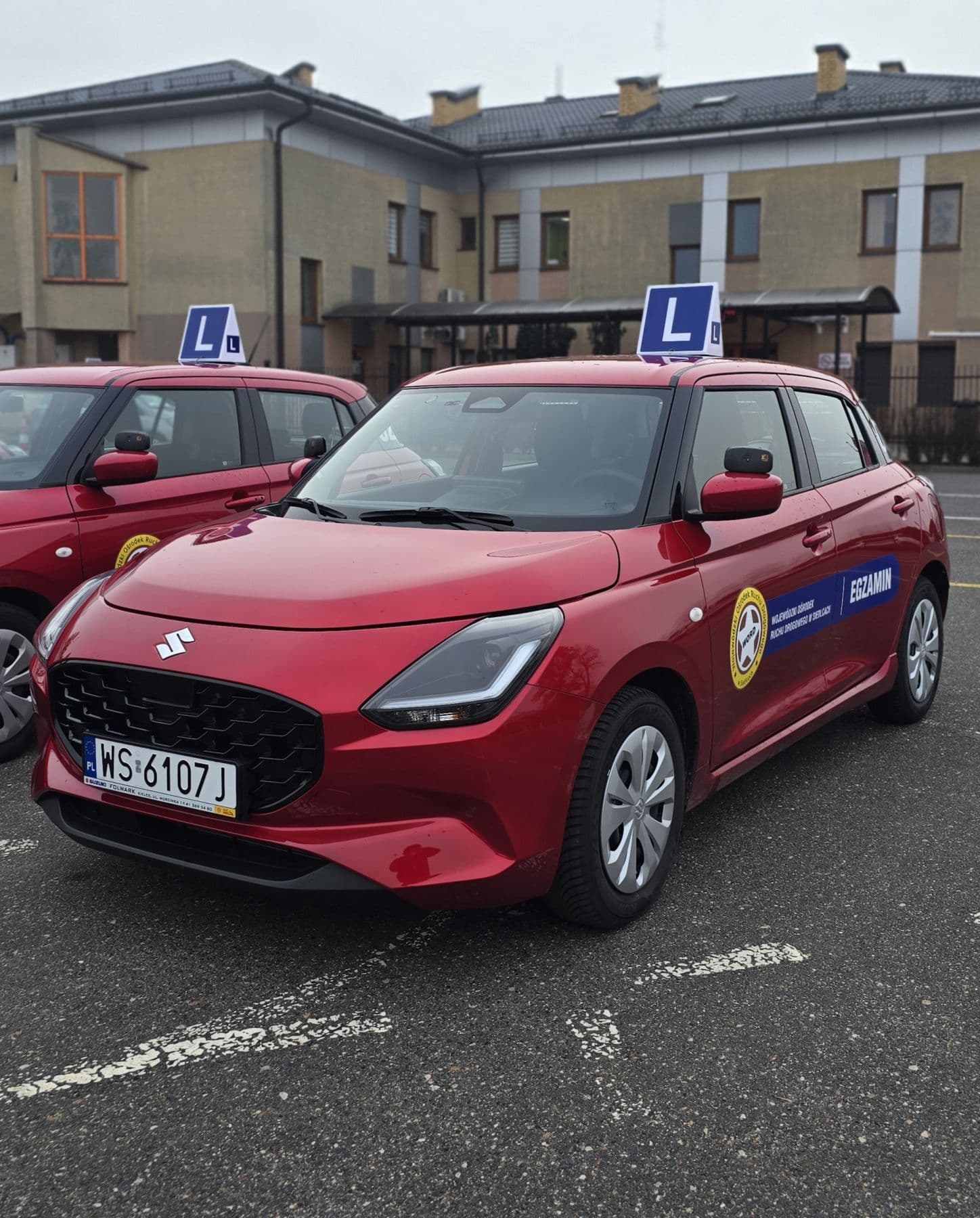 Dwa czerwone samochody szkoleniowe Suzuki Swift z tablicami „L” na dachach, zaparkowane obok siebie na placu manewrowym.